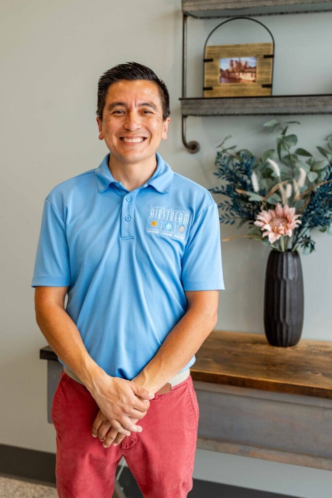 Smiling Airstream Plumbing, Heating & Air team member in a blue polo shirt, standing in an office or waiting area.