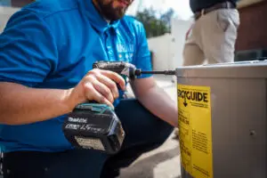 HVAC technician in a blue uniform using a drill on an outdoor air conditioning unit with an EnergyGuide sticker.