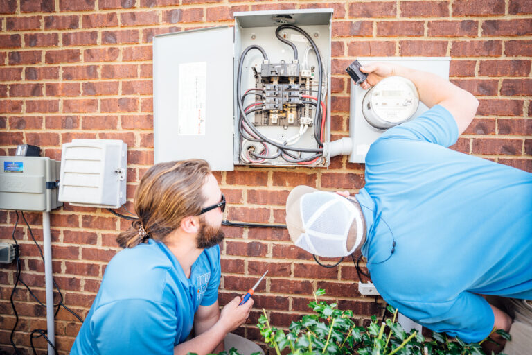 Two Airstream electricians inspecting and working on an outdoor electrical meter and service panel on a brick wall.