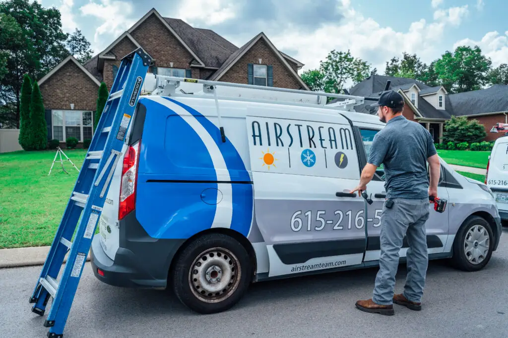 Airstream Plumbing, Heating & Air technician opening a branded service van door, with a ladder leaning against the back, outside a residential home.